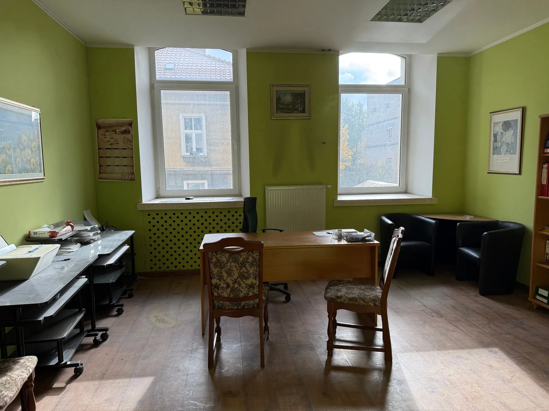 Wnętrze biura z jasnozielonymi ścianami i drewnianą podłogą. W centrum znajduje się drewniane biurko z dwoma krzesłami o tapicerowanych oparciach w kwiatowy wzór. Po lewej stronie stoi długi, szary stół roboczy z dokumentami i sprzętem biurowym. W tle widoczne są dwa okna z widokiem na sąsiednie budynki, biały grzejnik pod jednym z nich oraz dwa obrazy w ramach wiszące na ścianach. Po prawej stronie znajdują się dwa czarne fotele oraz regał z książkami.