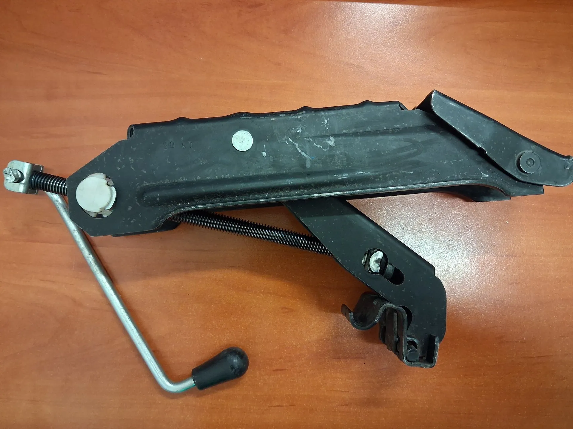 Czarna, metalowa część mechaniczna, prawdopodobnie mechanizm podnośnika szyby samochodowej, leżąca na brązowej, drewnianej powierzchni. Element składa się z głównej ramy, sprężyny oraz metalowej korby z czarną, plastikową rączką.