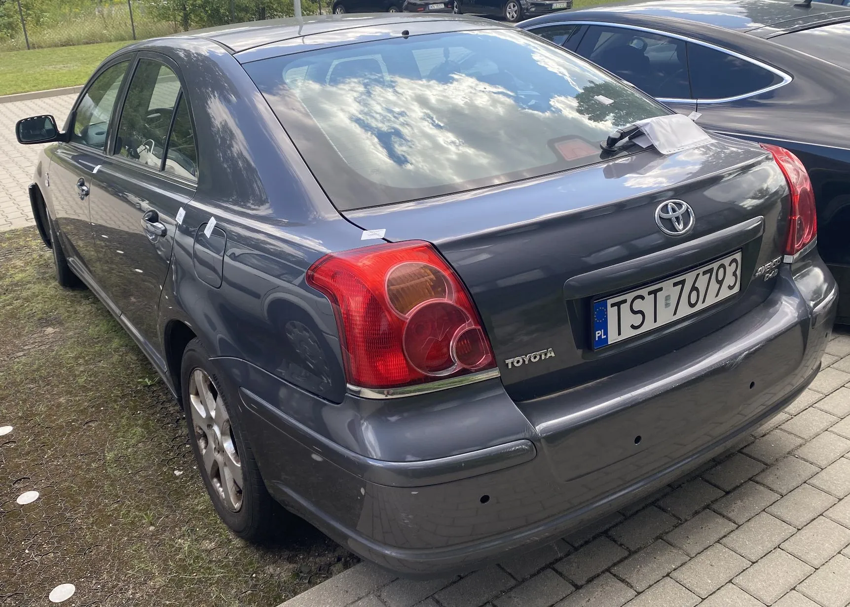 Tylny widok szarego samochodu marki Toyota z polską tablicą rejestracyjną o numerze TST 76793. Auto zaparkowane jest na kostce brukowej obok trawnika. Na klapie bagażnika widoczne jest logo Toyoty oraz napis TOYOTA.