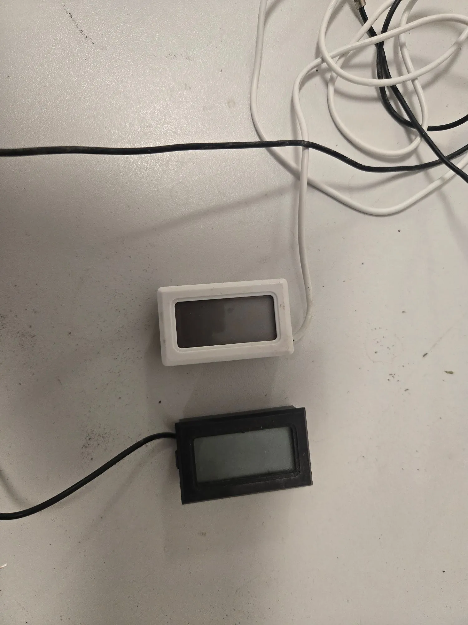 Dwa prostokątne urządzenia z wyświetlaczami LCD leżące na białej powierzchni. Górne urządzenie ma białą obudowę i jest podłączone białym kablem. Dolne urządzenie ma czarną obudowę i jest podłączone czarnym kablem. W górnej części obrazu widoczne są splątane białe i czarne przewody.