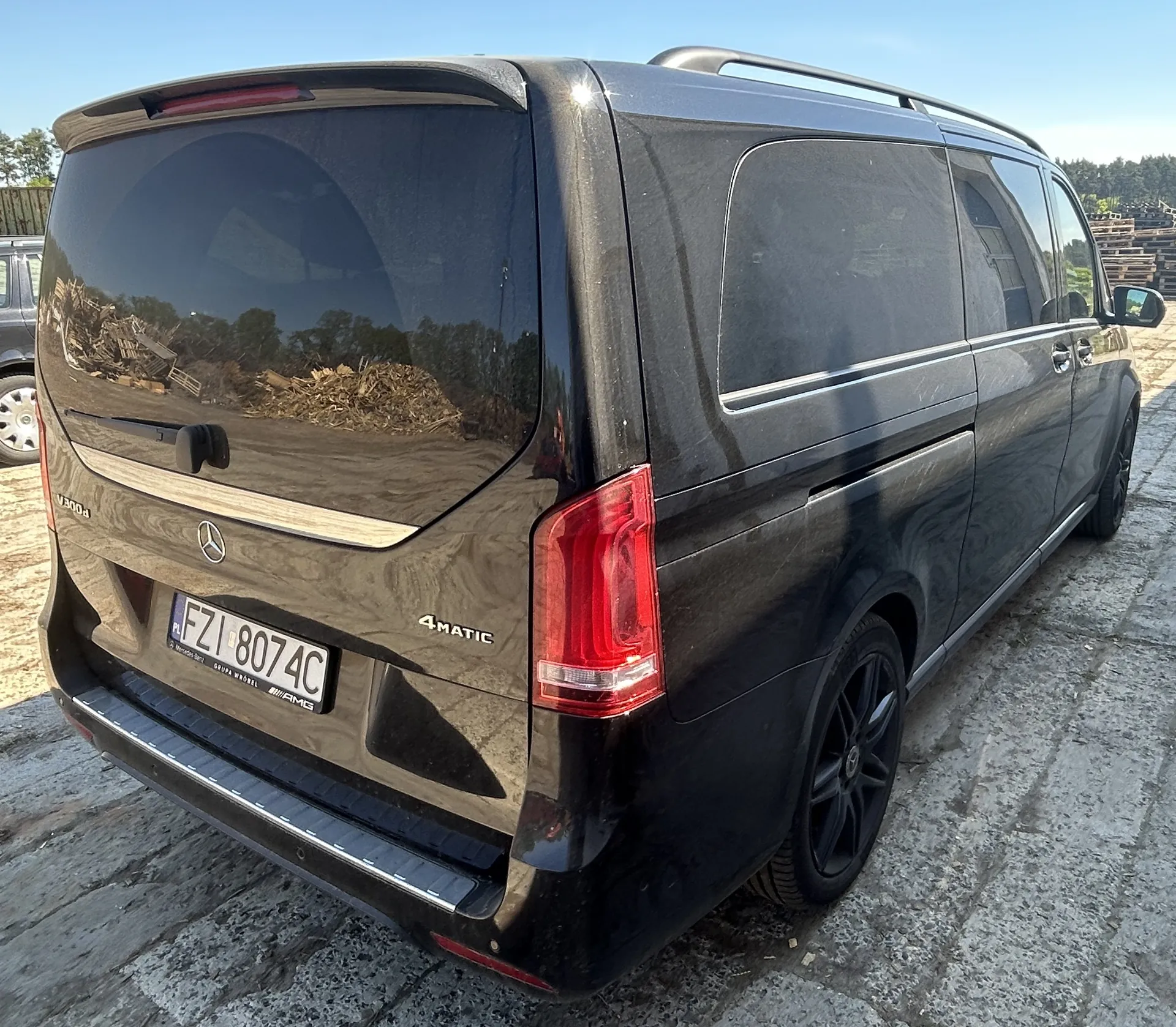 Czarny samochód Mercedes-Benz V300d z napędem 4MATIC, widok z tyłu i boku. Na tylnym zderzaku widoczny tabliczka rejestracyjna PL FZ1 8074C oraz logo Mercedes-Benz. W tle odbicie w szybie przedstawia stosy drewna i drzewa, a na lewo częściowo widoczny inny pojazd. Samochód stoi na brukowanej powierzchni pod jasnym niebem.