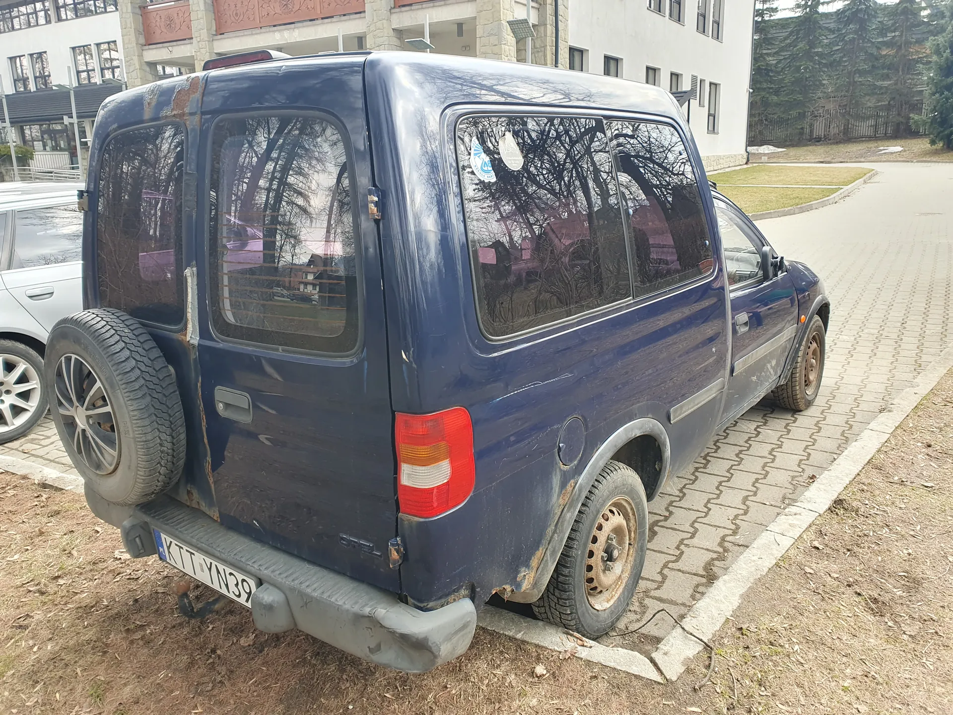 Niebieski furgon Opel zaparkowany na brukowanym placu. Widoczna tablica rejestracyjna KTT YN39, zapasowe koło na tylnej klapie, zarysowane reflektory i zanieczyszczenia na karoserii. W tle budynek z kamiennej elewacji, fragment srebrnego samochodu po lewej stronie oraz trawnik z drzewami. Na szybie tylnej naklejki, a na ziemi sucha trawa i liście.