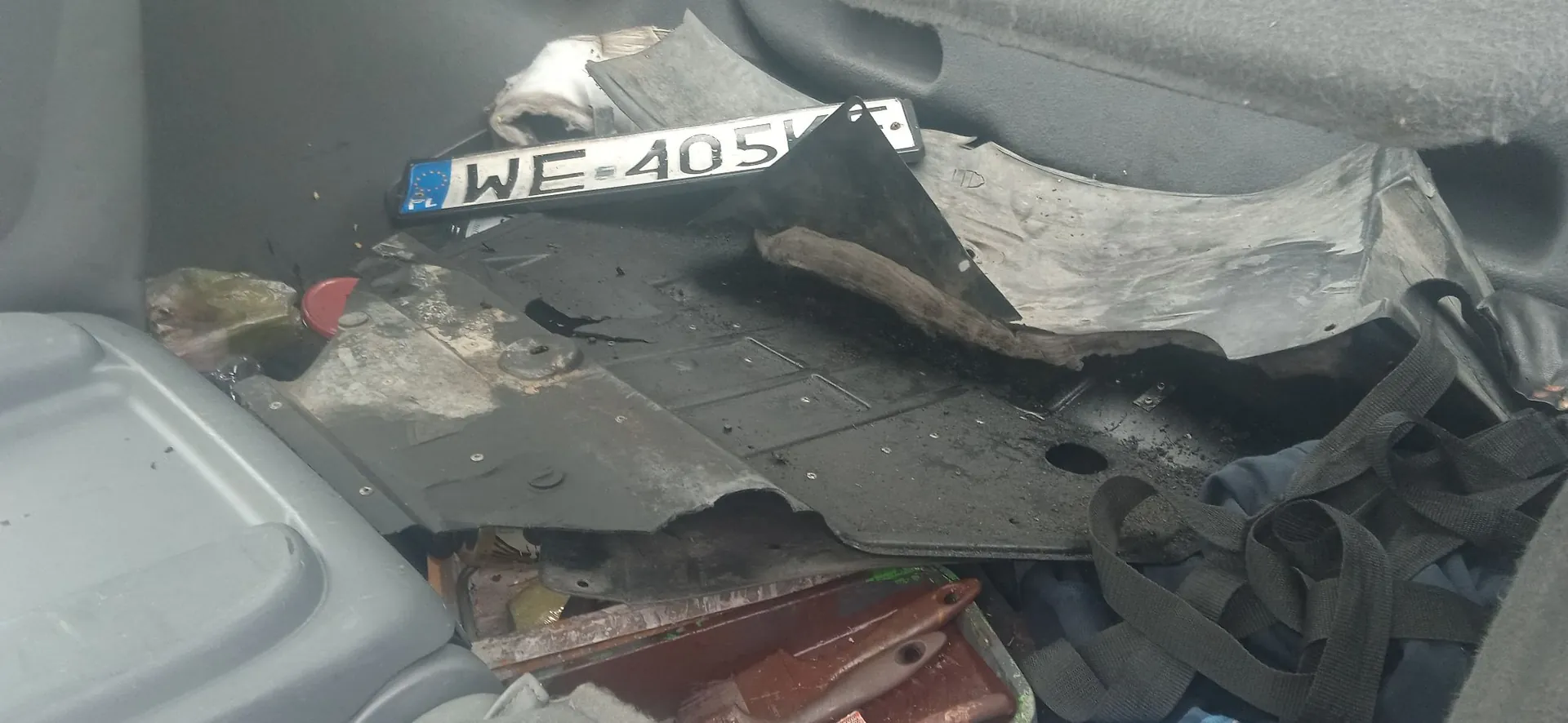 Wnętrze bagażnika samochodu wypełnione różnymi przedmiotami. Widoczna jest polska tablica rejestracyjna o numerze WE 405V, leżąca poziomo na czarnym, zabrudzonym materiale. Obok znajdują się fragmenty plastikowych osłon, szara tkanina oraz brązowy, metalowy element konstrukcyjny na dnie.
