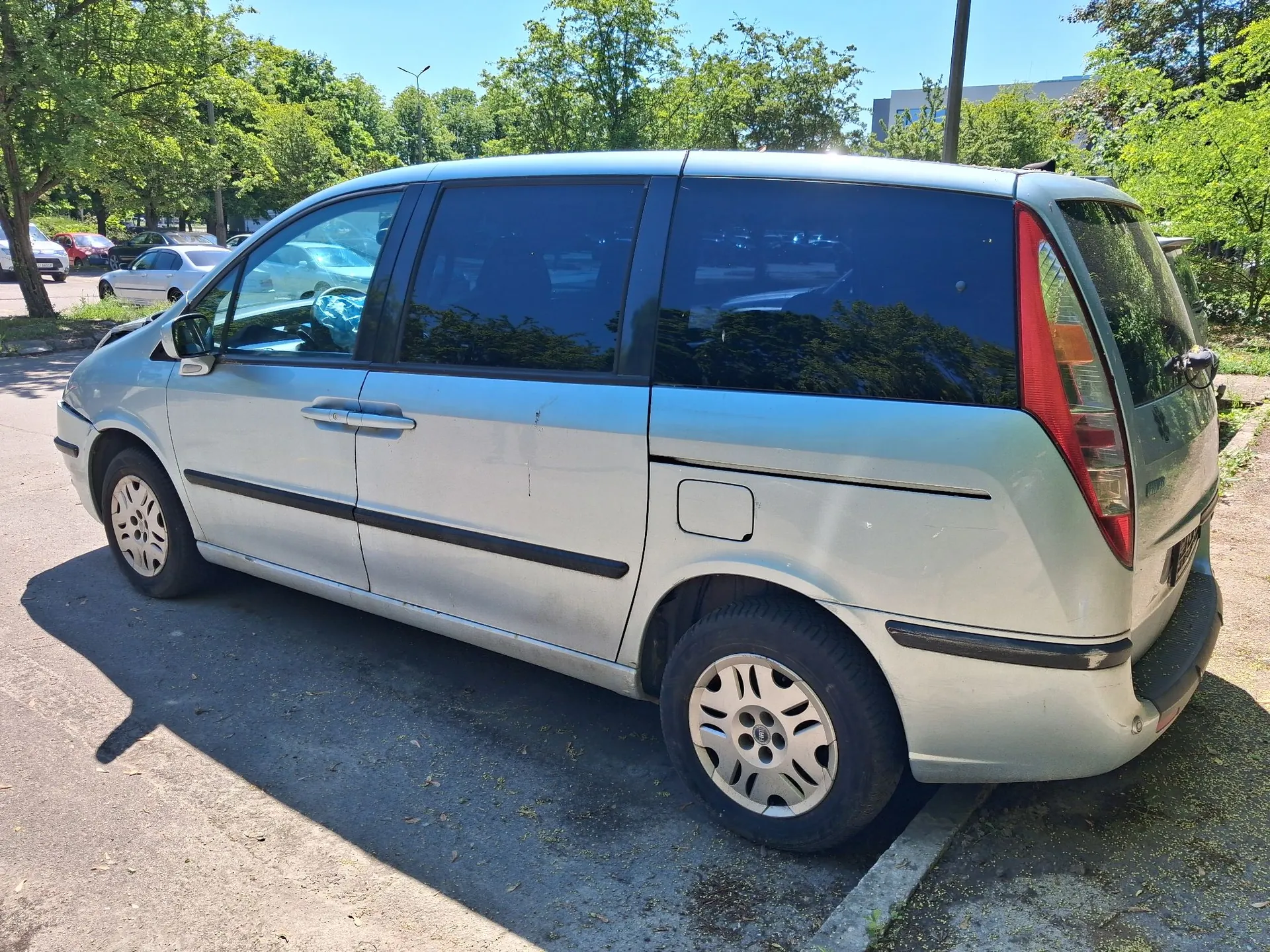 Srebrny samochód typu minivan zaparkowany na placu. Widok z boku i z tyłu pojazdu. W tle znajdują się zielone drzewa oraz inne zaparkowane samochody pod jasnym, słonecznym niebem.
