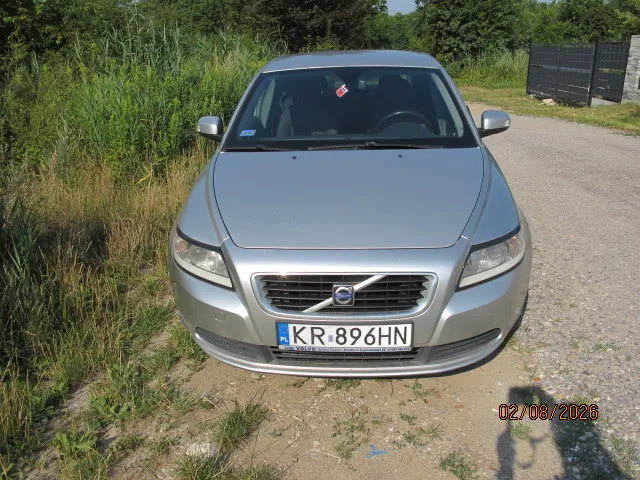 Srebrny samochód marki Volvo zaparkowany na piaszczystej drodze. Widoczny jest przód pojazdu z polską tablicą rejestracyjną o numerze KR 896HN. W tle znajduje się zielona roślinność, krzewy oraz czarny metalowy płot po prawej stronie. W prawym dolnym rogu zdjęcia widnieje data 02/08/2026.
