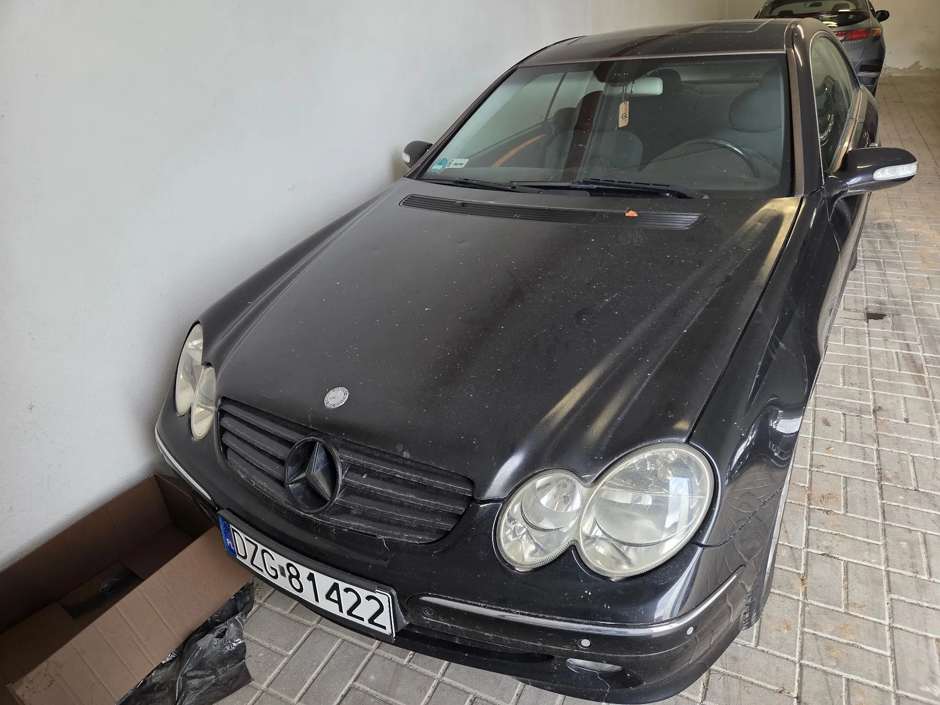 Czarny samochód marki Mercedes-Benz zaparkowany w garażu na kostce brukowej. Pojazd jest zakurzony i posiada polską tablicę rejestracyjną o numerze DZG 81422. Obok samochodu, po lewej stronie, widoczny jest fragment kartonowego pudełka.