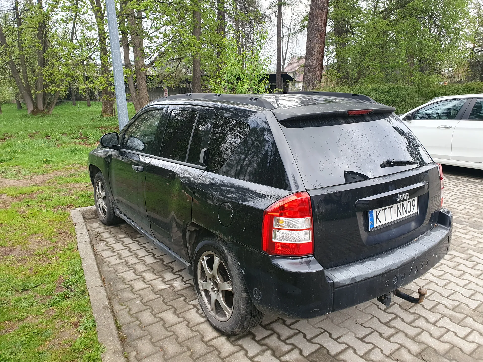 Czarny SUV Jeep Compass zaparkowany na brukowanej powierzchni, widok od tyłu. Widoczna tablica rejestracyjna z napisem KTT NN09 oraz oznaczeniem PL po lewej stronie. Na bagażniku widnieje logo Jeep i napis Compass. Po prawej stronie częściowo widoczny biały samochód. Tło stanowią drzewa, zielona trawa i domy w oddali. Samochód ma mokrą karoserię, co sugeruje niedawny deszcz.