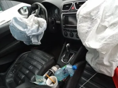 Wnętrze samochodu z rozwiniętymi poduszkami powietrznymi. Widoczne są rozwinięta poduszka kierowcy (niebiesko-biała) i pasażera (biała). Deska rozdzielcza z kierownicą, systemem audio i przyciskami. W centralnej konsoli dźwignia zmiany biegów. Na siedzeniu pasażera butelka wody, kubek z niedopałkiem papierosa oraz inne przedmioty. Czarne skórzane fotele. Wnętrze w stanie po wypadku.