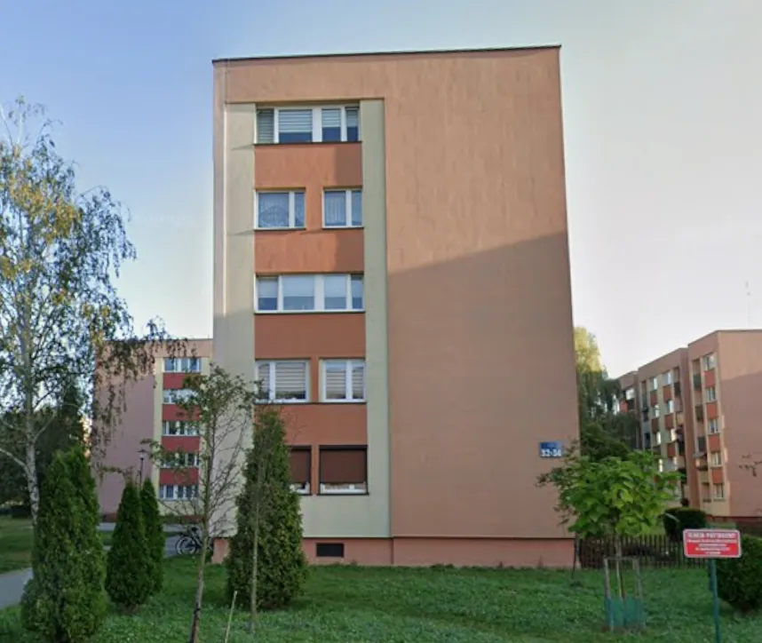 Wielopiętrowy budynek mieszkalny o beżowej i brązowej elewacji. Budynek posiada rzędy okien z białymi ramami i roletami. Przed budynkiem znajduje się trawnik oraz kilka zielonych, stożkowatych krzewów i drzew. Po prawej stronie widoczna jest czerwona tabliczka informacyjna na słupku oraz fragment innego, podobnego budynku.