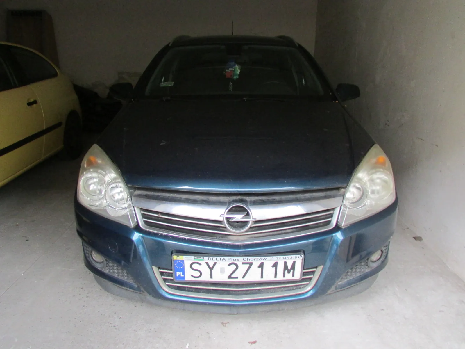 Widok z przodu ciemnoniebieskiego samochodu marki Opel zaparkowanego w garażu. Obok, po lewej stronie, widoczny jest fragment żółtego samochodu. Samochód posiada polską tablicę rejestracyjną o numerze SY 2711M. Nad numerem rejestracyjnym znajduje się napis: DELTA Plus Chorzów 32 246 346.
