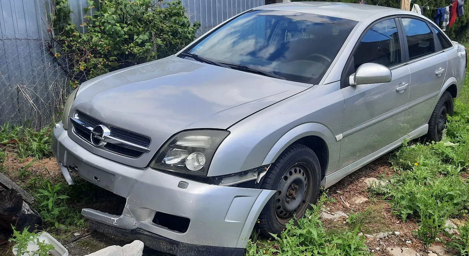 Srebrny samochód osobowy marki Opel zaparkowany na trawiastym terenie obok metalowego ogrodzenia. Pojazd jest uszkodzony: przedni zderzak jest częściowo oderwany i pęknięty, a maska nie domyka się szczelnie. Auto stoi na czarnych felgach stalowych.