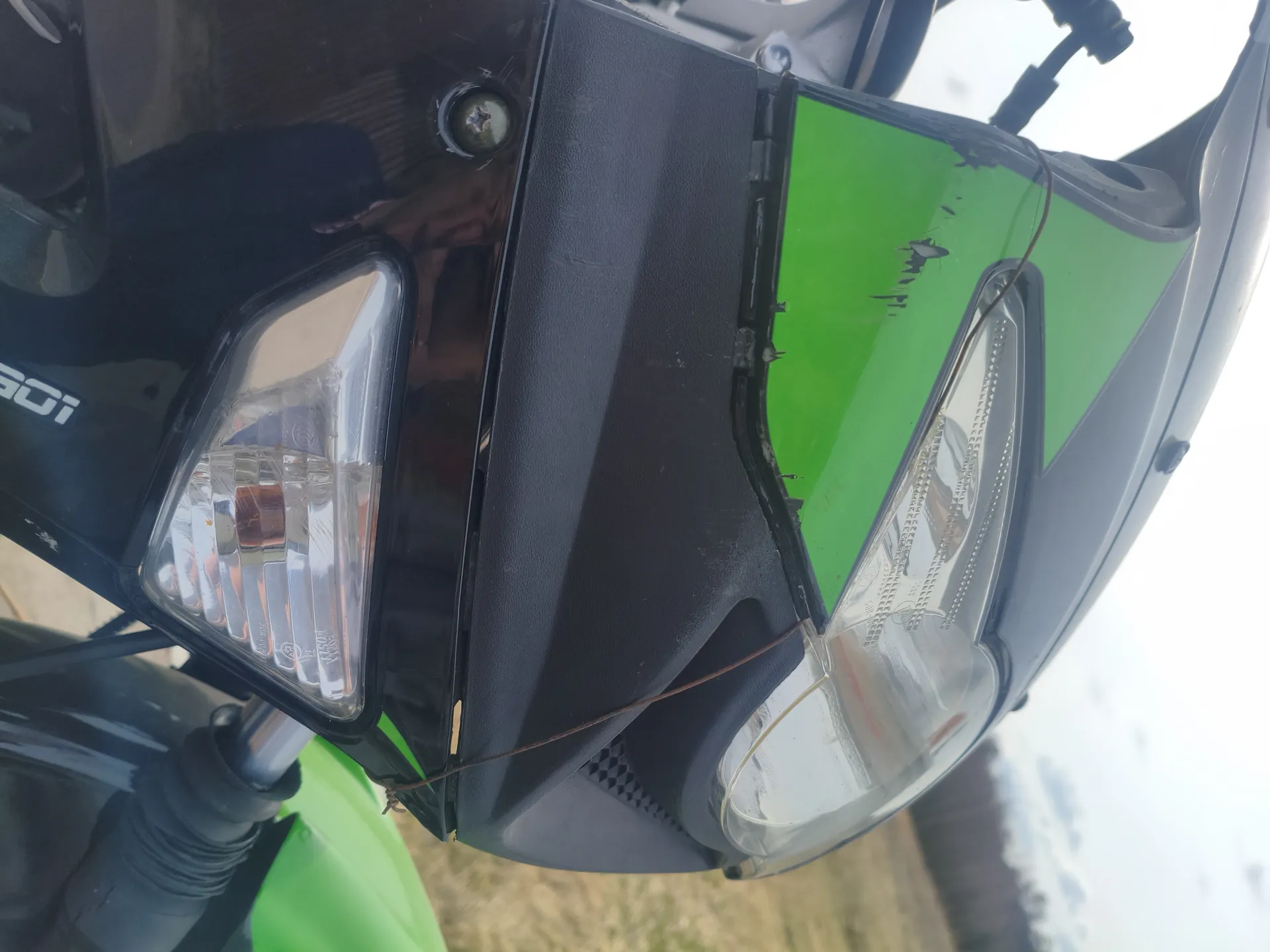 Zbliżenie na przednią część motocykla z zielonymi i czarnymi elementami karoserii, reflektorem głównym oraz dodatkowym światłem. Widoczne są zadrapania na zielonym panelu i cienki drut przymocowany do części przedniej. W tle rozległe pole i drzewa. Na czarnej powierzchni widoczny fragment napisu '100i'.