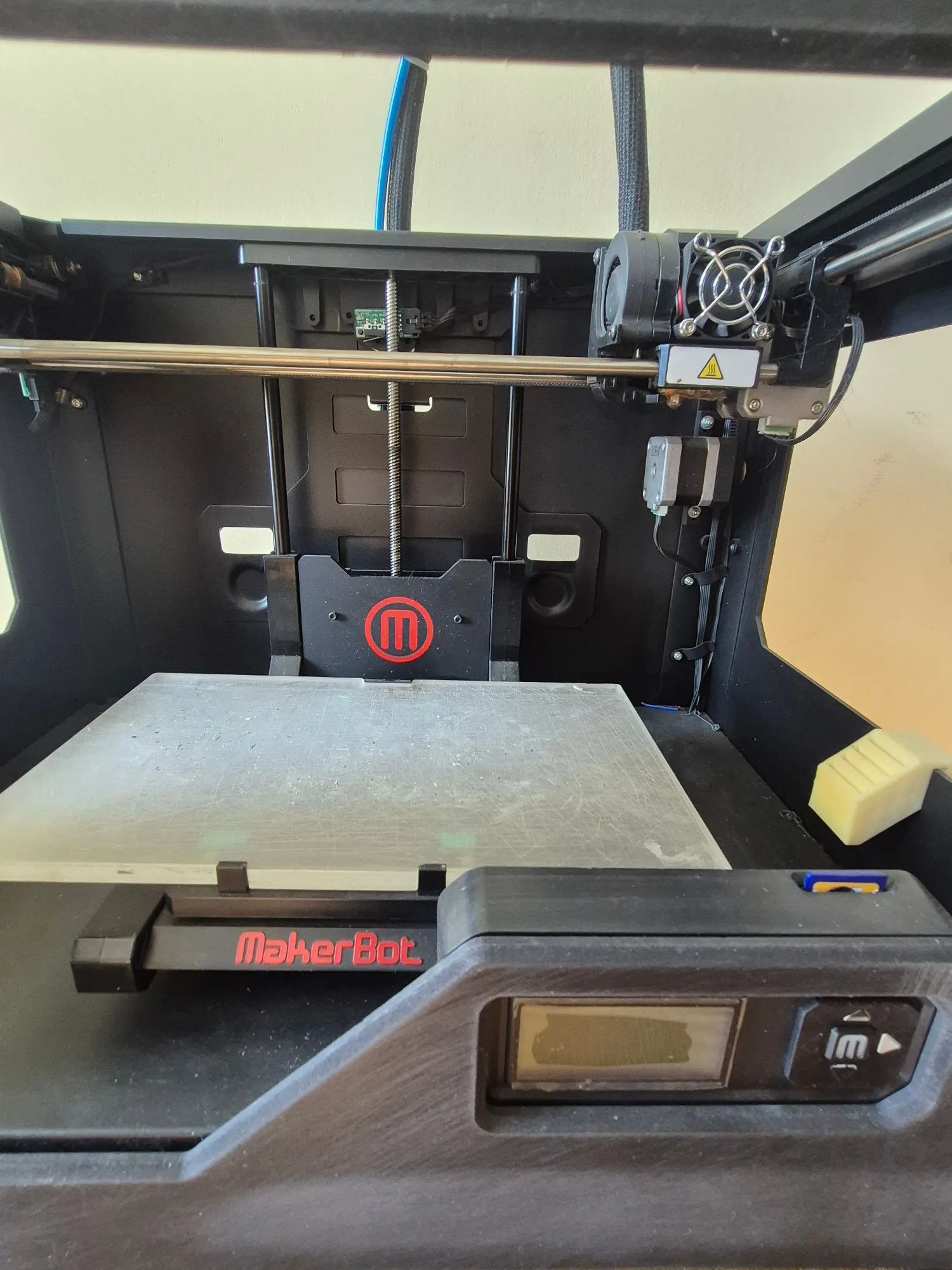 Zdjęcie wnętrza drukarki 3D marki MakerBot. Widoczny jest stół roboczy, oś X z głowicą drukującą oraz czarna obudowa z czerwonym logo MakerBot w kształcie litery 'm' w okręgu. Na przednim panelu znajduje się napis 'MakerBot' oraz mały ekran LCD z przyciskami sterującymi.