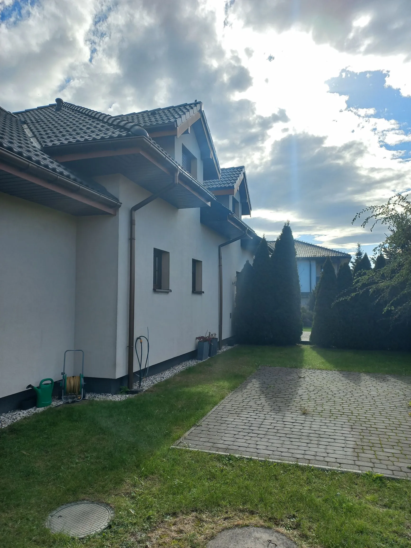 Widok z boku na biały, dwukondygnacyjny dom z ciemnoszarym, spadzistym dachem. Przy ścianie budynku znajduje się rynna, a obok niej zielona konewka i zwinięty wąż ogrodowy na stojaku. Przed domem rozciąga się trawnik z wybrukowanym, prostokątnym podjazdem oraz rzędem wysokich, smukłych tuj. W tle widoczne jest jasne niebo z rozproszonymi chmurami i słońcem przebijającym się przez nie.