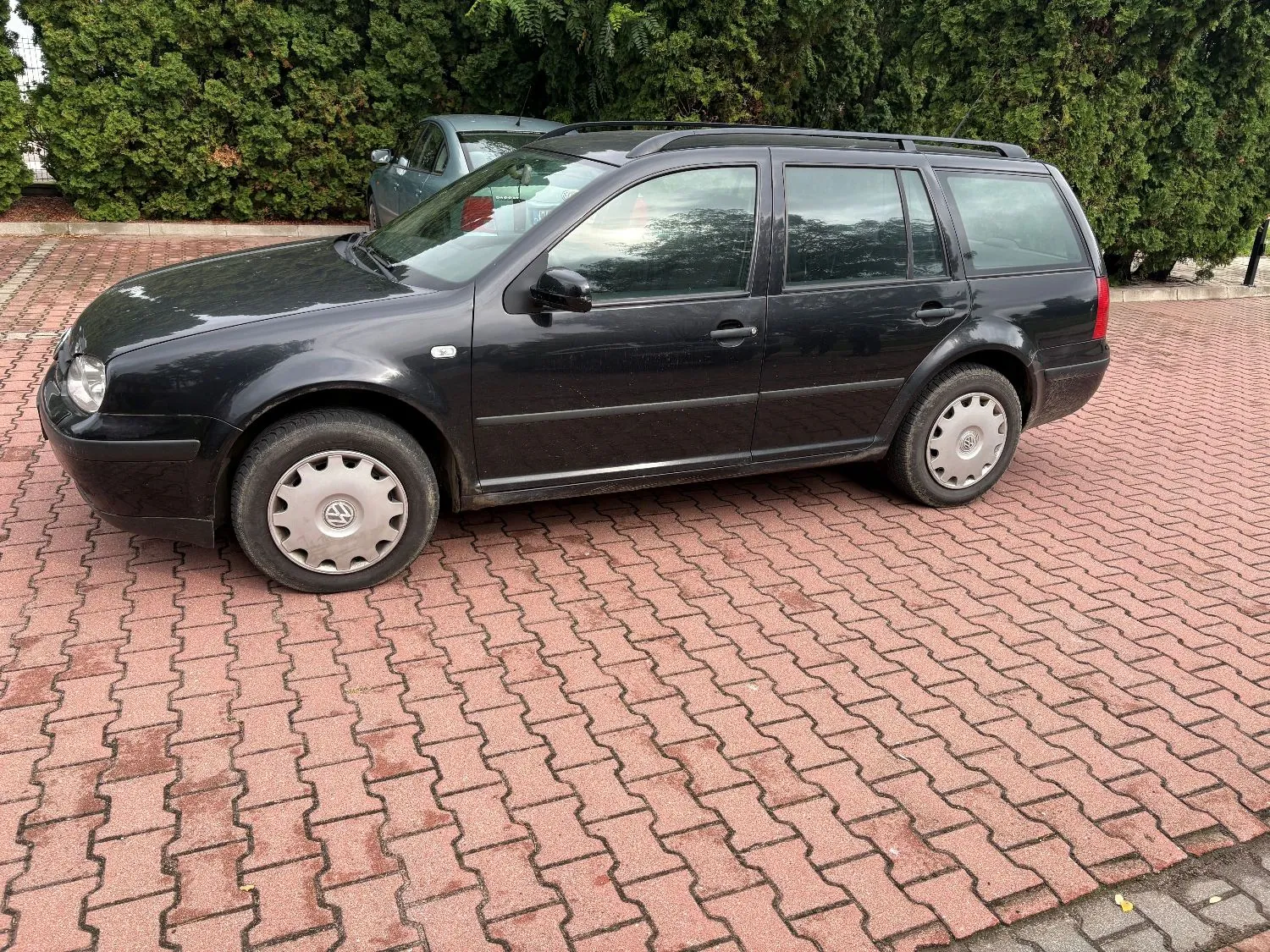 Czarny samochód marki Volkswagen Golf w wersji kombi zaparkowany bokiem na czerwonej kostce brukowej. W tle widoczny jest inny, jasnoniebieski samochód oraz gęsty, zielony żywopłot.