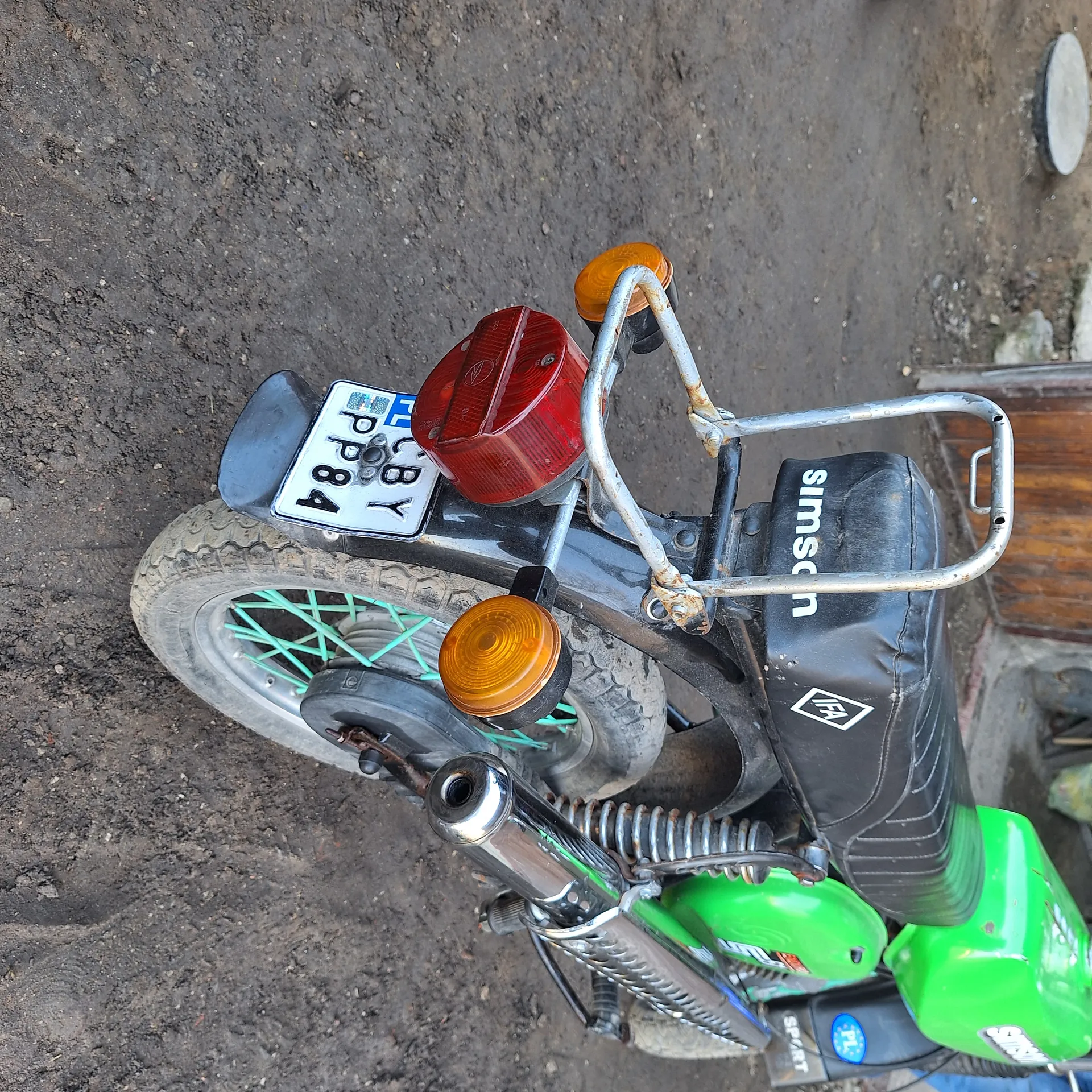 Z tyłu motocykla marki Simson widoczne są zielone elementy, czarne siedzenie z napisem SIMSON, tablica rejestracyjna z napisem CBY PP84, czerwone światło tylne oraz pomarańczowe kierunkowskazy. Koło tylne z zielonymi promieniami i brudną oponą. Tło stanowi brudna, ziemista powierzchnia.