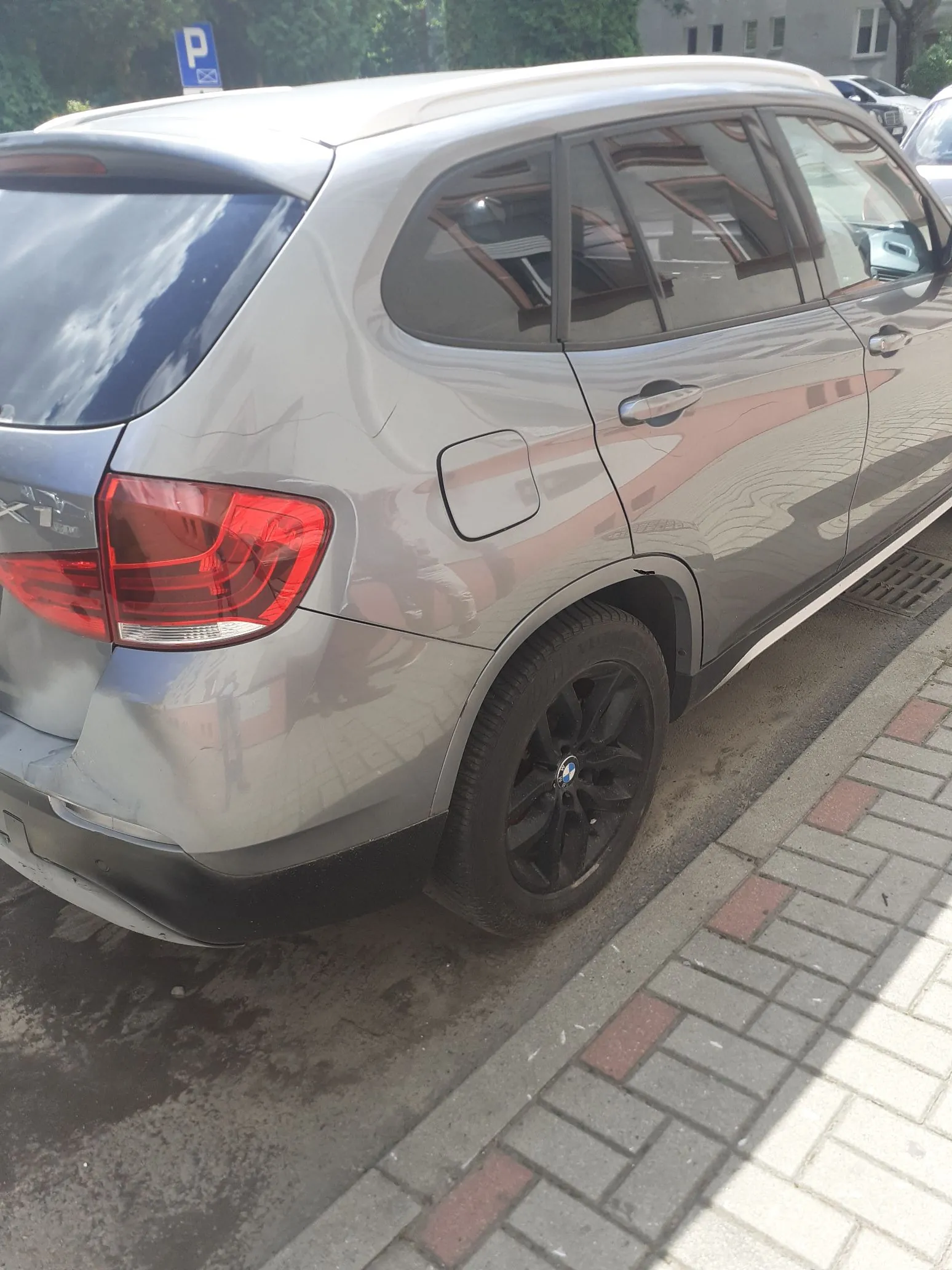 Srebrny samochód marki BMW typu SUV zaparkowany przy krawężniku na chodniku z kostki brukowej. Widoczny jest tył i prawy bok pojazdu, w tym tylna lampa, czarna felga oraz wlew paliwa. W tle widać zielone krzewy oraz niebieski znak parkingowy z literą P.