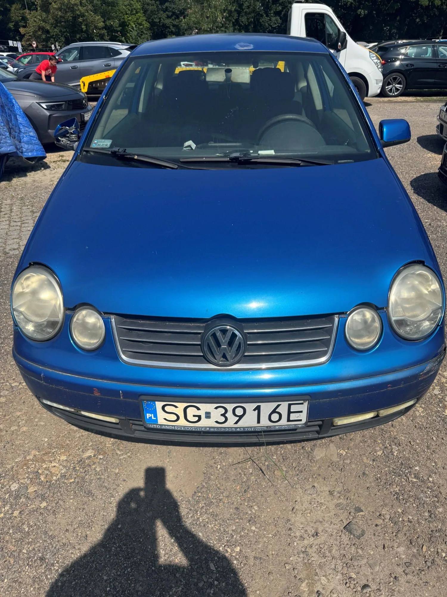 Zdjęcie przodu niebieskiego samochodu marki Volkswagen. Pojazd posiada polską tablicę rejestracyjną o numerze SG 3916E. W tle widoczne są inne samochody zaparkowane na placu oraz drzewa.