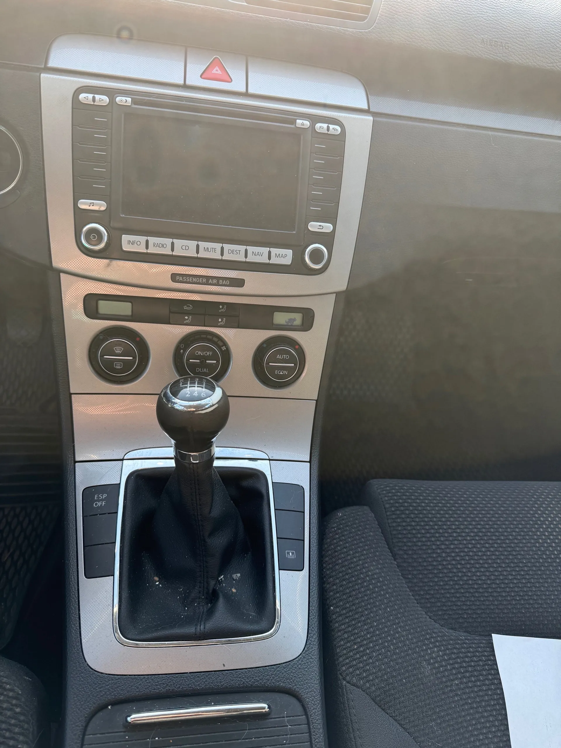 Konsola środkowa samochodu z czarną dźwignią zmiany biegów oznaczoną pozycjami 1-6 i R. Powyżej ekran dotykowy systemu rozrywki z przyciskami: INFO, RADIO, CD, MUTE, DEST, NAV, MAP. Poniżej ekranu napis 'PASSENGER AIR BAG'. Trzy okrągłe pokrętła klimatyzacji: lewe z ikoną wentylatora, środkowe z napisem 'ON/OFF DUAL', prawe z napisem 'AUTO ECON'. Po lewej stronie przycisk 'ESP OFF'. Widoczne fragmenty czarnych siedzeń i fragment deski rozdzielczej w kolorze szarym.