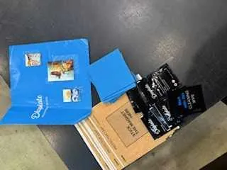 Niebieska teczka z logo DHL i kilkoma naklejkami, w tym z widocznym obrazkiem, obok brązowego kartonu oraz czarnych paczek z logo DHL. Wszystkie elementy leżą na ciemnej powierzchni.