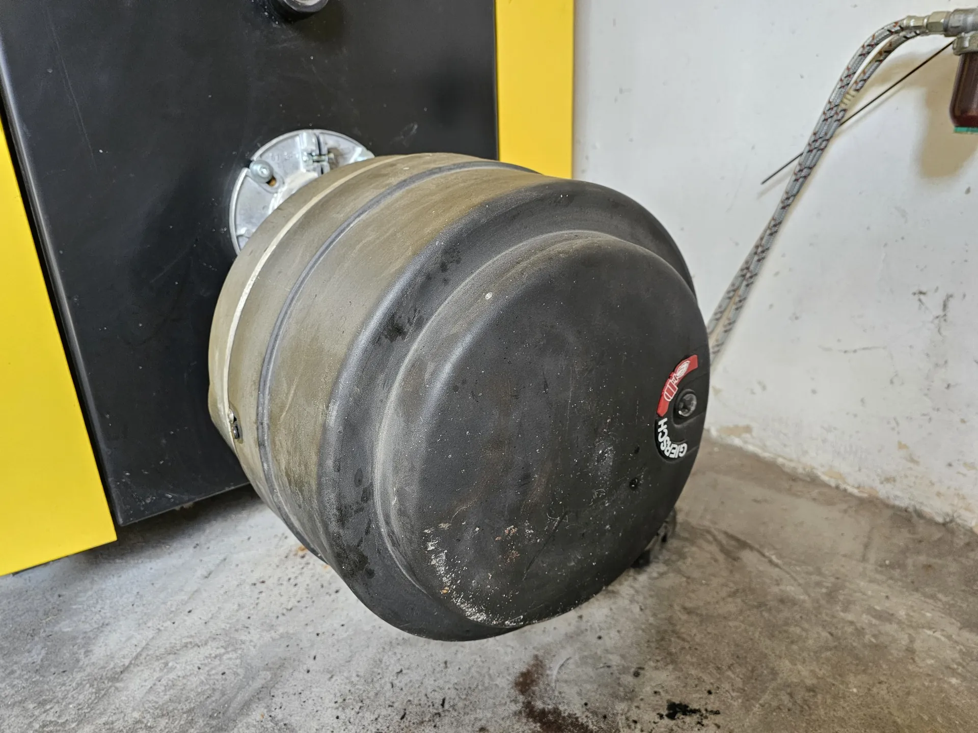 Zbliżenie na czarny, cylindryczny element mechaniczny, prawdopodobnie siłownik lub zbiornik, zamontowany do czarnej obudowy urządzenia. Przedmiot jest zakurzony i posiada niewielki czerwono-biały napis z logo oraz słowem 'Hesch'. W tle widoczna jest biała ściana oraz szary, betonowy podłoga.