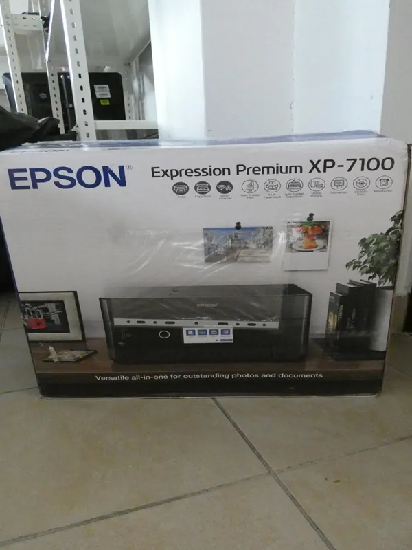 Pudełko drukarki Epson Expression Premium XP-7100. Na białym opakowaniu widoczny niebieski napis EPSON, model drukarki oraz ikony funkcji. Poniżej umieszczono zdjęcie czarnej drukarki, dwa przykładowe zdjęcia wydruków (scena uliczna i talerz z jedzeniem) oraz napis: Versatile all-in-one for outstanding photos and documents. Tło przedstawia pomieszczenie z półkami i podłogą z kafli.