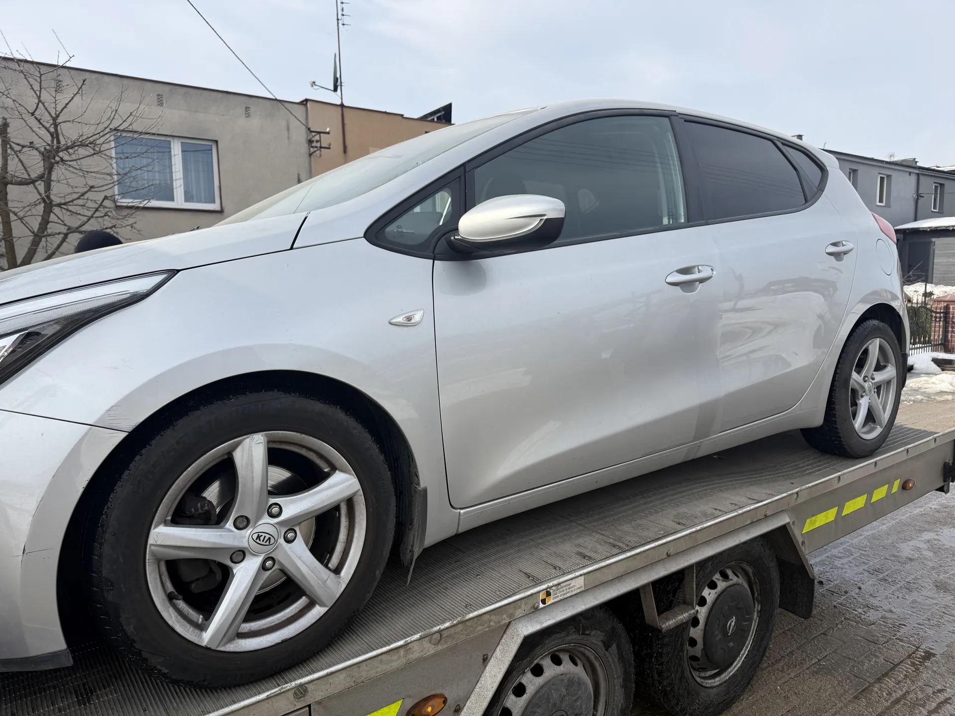 Srebrny samochód marki Kia na płycie holownika. Widoczne są felgi z logo KIA, boczne lusterko, drzwi i przednia część pojazdu. Holownik ma żółte znakowanie na krawędzi. W tle budynki mieszkalne, bezlistne drzewo, resztki śniegu na ziemi oraz pochmurne niebo.