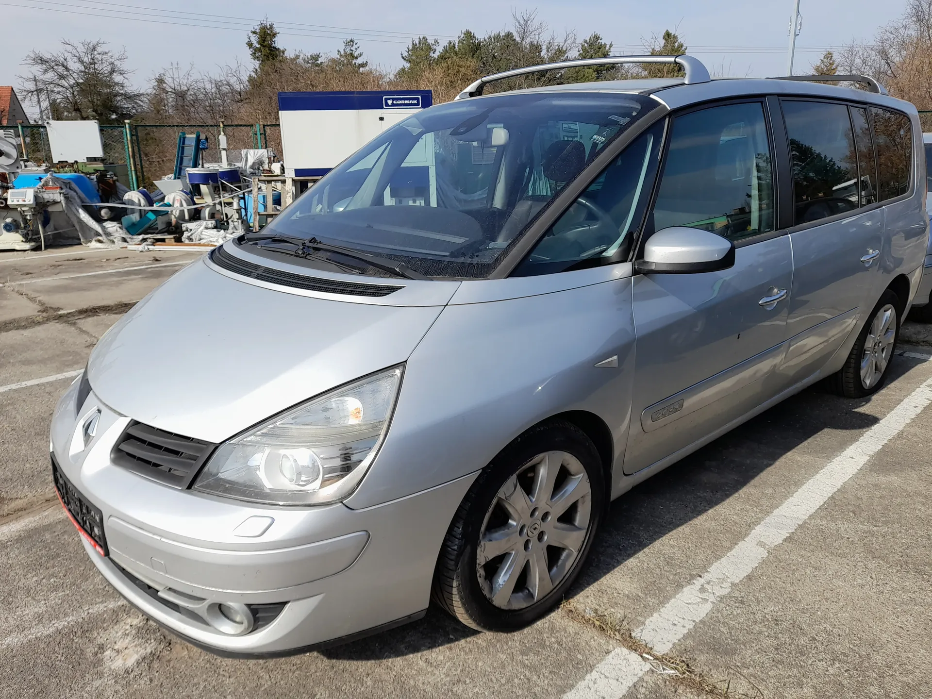 Srebrny samochód typu minivan z przymocowanymi na dachu białymi bagażnikami, zaparkowany na asfaltowym placu parkingowym z białymi liniami. W tle widoczne są przemysłowe urządzenia, niebiesko-biały kontener z napisem CORMAK, ogrodzenie, drzewa oraz jasne niebo. Samochód posiada czarne felgi, przednie reflektory i chromowaną atrapę chłodnicy. Na pierwszym planie widoczne są fragmenty asfaltu z białymi liniami parkingu.