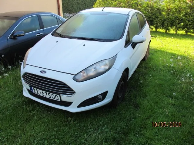Biały samochód marki Ford z tablicą rejestracyjną KK-67500 zaparkowany na trawie. W tle widoczny czarny samochód, zielone krzewy i fragment budynku. Na trawie rosną białe kwiaty. W prawym dolnym rogu obrazu znajduje się data 19/05/2026.