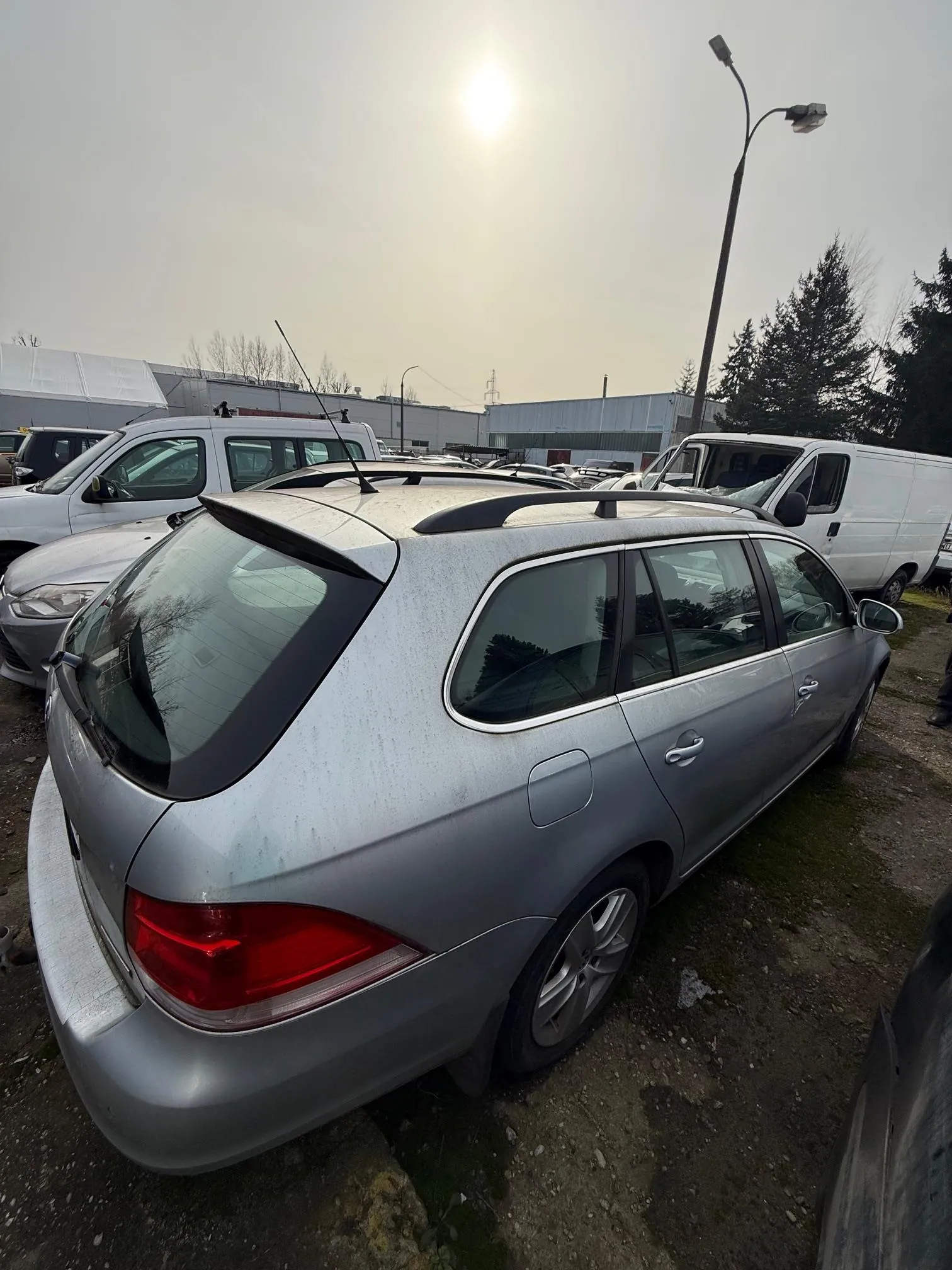 Srebrny samochód typu kombi zaparkowany na otwartym placu. W tle widoczne są inne pojazdy, w tym białe furgony, budynki przemysłowe, drzewa oraz słoneczna pogoda. Na pierwszym planie samochód ma czarne dachówki, czerwone światła tylnego oraz metalowe felgi. Podłoga jest nierówna, z widoczną ziemią i plamami mchu. W oddali znajduje się latarnia uliczna i słoneczne niebo.