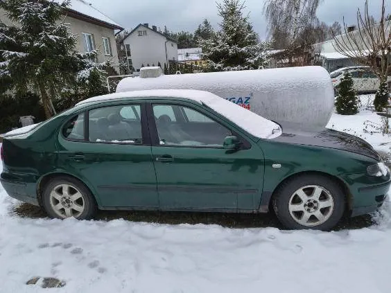 Zielony samochód osobowy typu sedan pokryty śniegiem, zaparkowany na śnieżnej powierzchni. Za nim duży biały zbiornik z częściowo widocznym napisem "GAZ". W tle domy, drzewa pokryte śniegiem oraz inne pojazdy. Niebo jest zachmurzone.