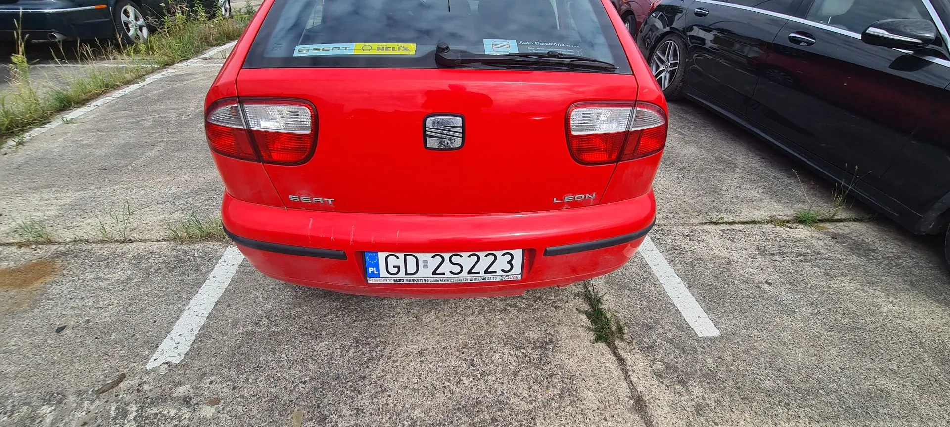 Tylny widok czerwonego samochodu marki Seat Leon zaparkowanego na betonowym parkingu. Na klapie bagażnika widoczne jest logo marki, napis SEAT po lewej stronie oraz LÉON po prawej. Samochód posiada polską tablicę rejestracyjną o numerze GD 2S223. W tylnej szybie znajdują się naklejki, w tym logo SEAT i HELIX oraz informacja o Auto Barcelona. Po prawej stronie zdjęcia widoczny jest fragment czarnego samochodu.