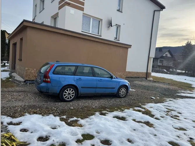 Niebieski samochód typu kombi zaparkowany na żwirowym podjeździe przed dwupiętrowym domem o białych i brązowych ścianach. Na ziemi widoczne są resztki śniegu oraz zielona trawa. W tle domy, wzgórza i częściowo zachmurzone niebo. Na ścianie budynku znajduje się antena satelitarna.