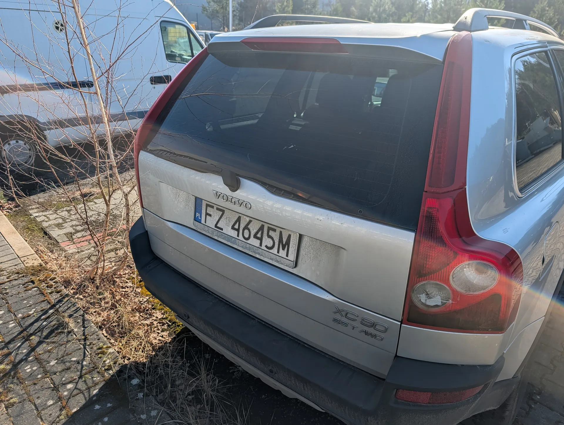 Z tyłu srebrny samochód Volvo XC 90 2.5 T AWD z tablicą rejestracyjną FZ 4645M (PL). W tle biały furgon, suche krzewy i brukowana powierzchnia. Na samochodzie widoczne są czerwone światła tylnego oraz napisy 'VOLVO' i 'XC 90 2.5 T AWD'.