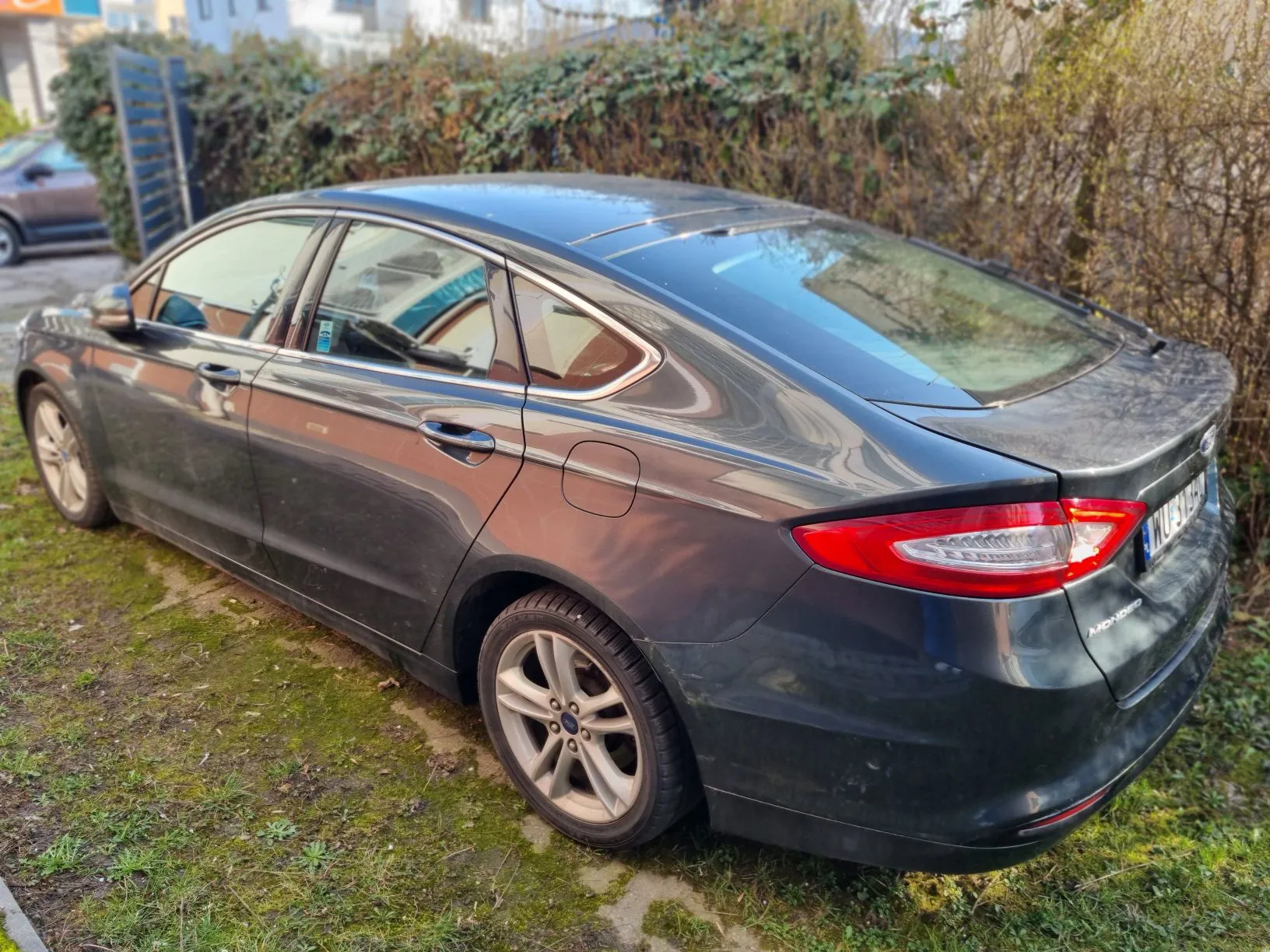 Ciemnoszary samochód Ford Mondeo zaparkowany na trawie, widok z tyłu i boku. Pojazd posiada dach panoramiczny, felgi aluminiowe oraz widoczną tablicę rejestracyjną WU 194. W tle krzewy, niebieskie ogrodzenie oraz częściowo widoczny inny samochód. Scena na zewnątrz w słoneczny dzień.
