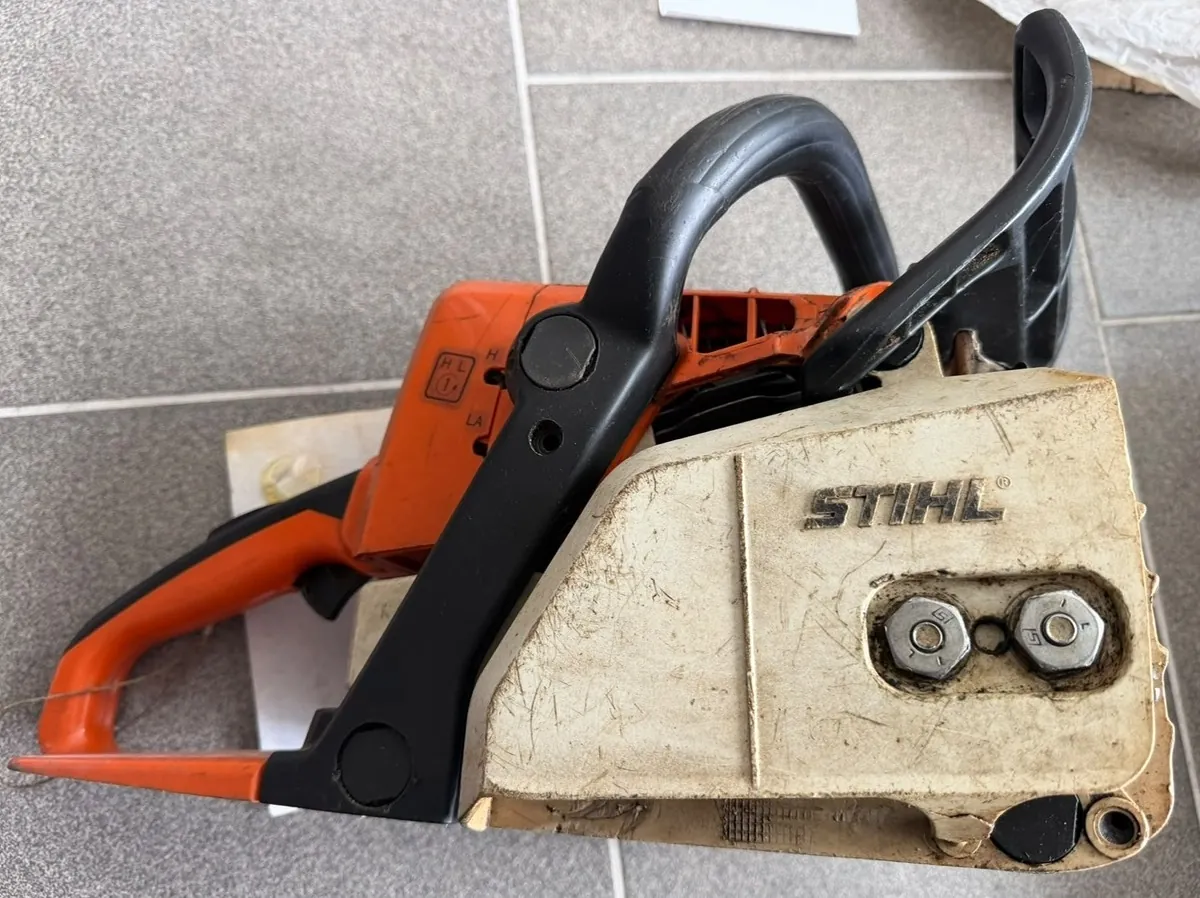 Zdjęcie przedstawiające piłę spalinową marki Stihl, leżącą na szarych płytkach ceramicznych. Urządzenie ma pomarańczową obudowę silnika i biały element osłony z widocznym napisem 'STIHL'. Widoczny jest czarny uchwyt oraz dwie metalowe śruby mocujące w białej części obudowy.