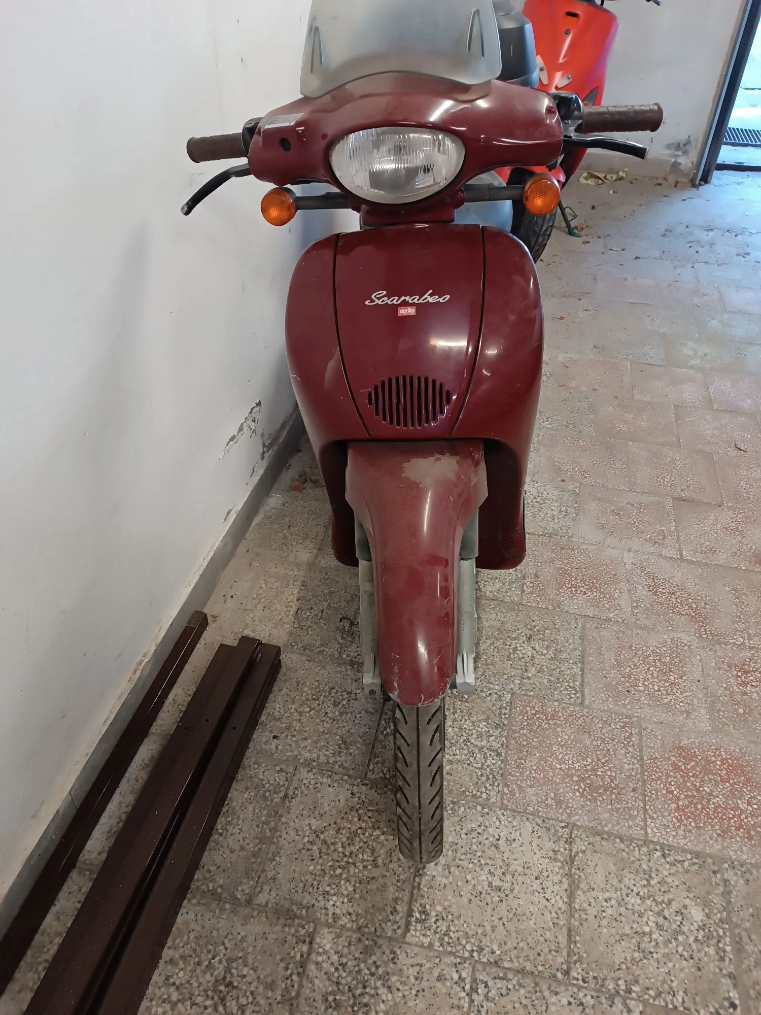 Widok z przodu na bordowy skuter marki Aprilia z napisem 'Scarabeo' na przedniej owiewce. Pojazd posiada pojedynczy reflektor, pomarańczowe kierunkowskazy i szary błotnik. Skuter stoi zaparkowany na jasnej, płytkowej podłodze przy białej ścianie.