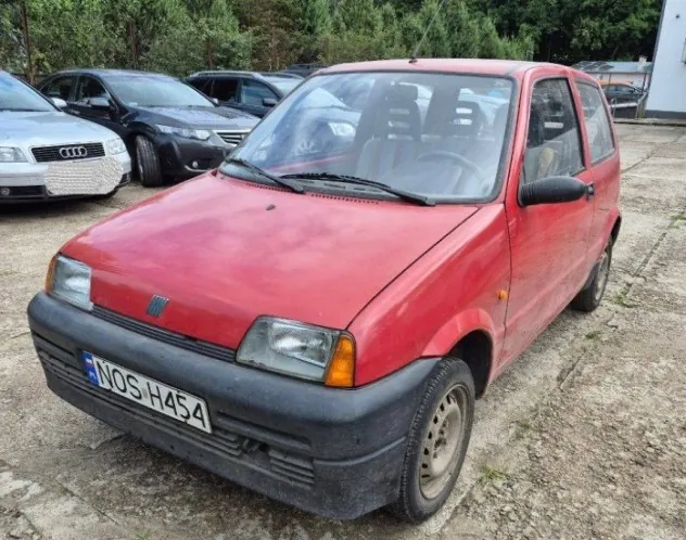 Czerwony, trzydrzwiowy samochód osobowy marki Fiat, zaparkowany na nieutwardzonym placu. Pojazd ma czarny plastikowy zderzak przedni z tablicą rejestracyjną o numerze NOS H454. W tle widoczne są inne samochody, w tym biały Audi oraz czarny samochód, a za nimi pas zieleni z drzewami i krzewami.