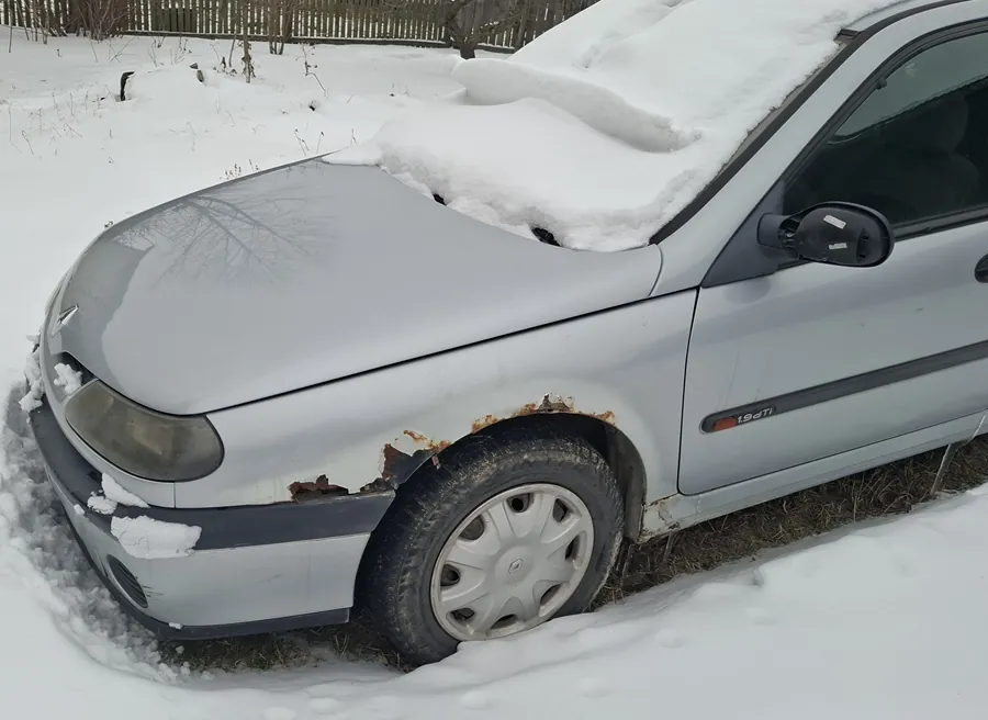 Srebrny samochód osobowy zaparkowany na śniegu. Na masce i dachu widoczna warstwa śniegu. Przednia lewa część nadkola pokryta rdzą. Na boku nadwozia napis "1.9 dTi". Tło: zasneżony teren, drewniane ogrodzenie i nagie drzewa. Opona przedniego koła jest zużyta, a na masce odbija się sylwetka drzewa.