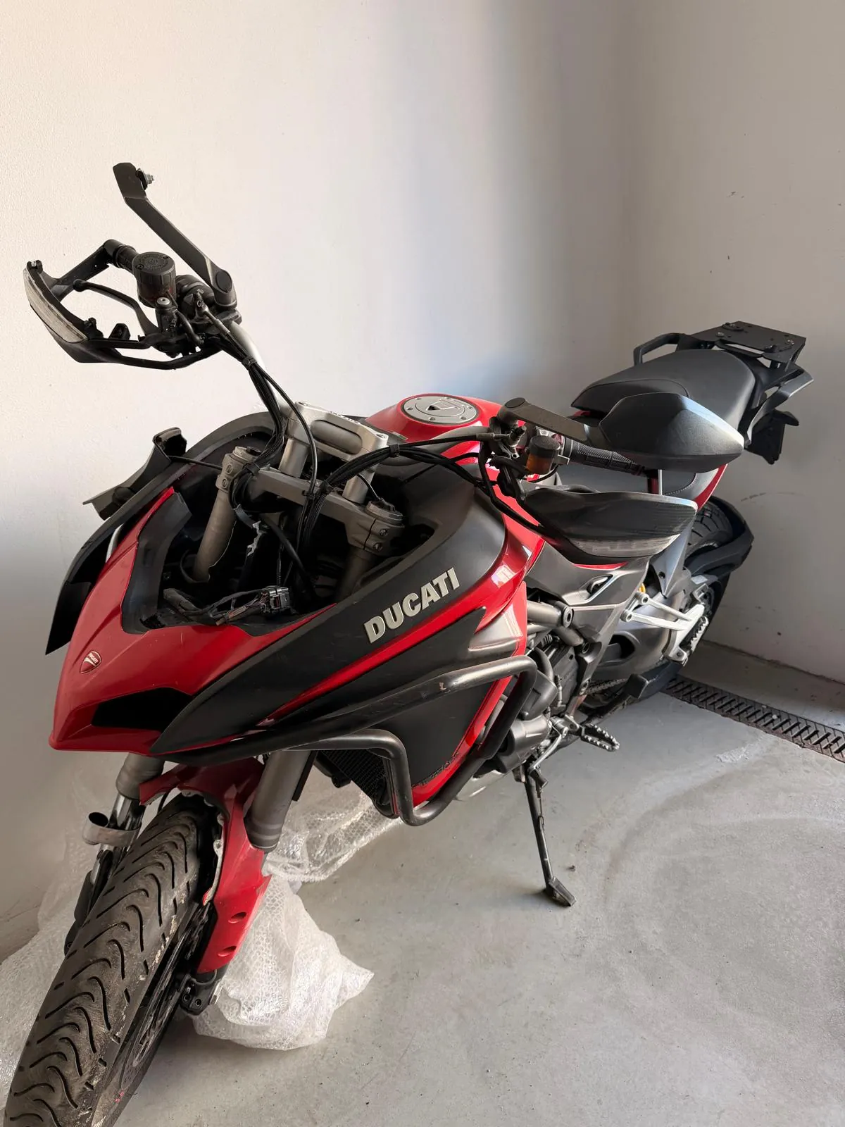 Czerwony motocykl marki Ducati zaparkowany w białym pomieszczeniu. Motocykl jest ustawiony bokiem, widoczny jest napis 'DUCATI' na owiewce. Pojazd stoi na bocznej stopce, a pod przednim kołem znajduje się biała folia bąbelkowa.