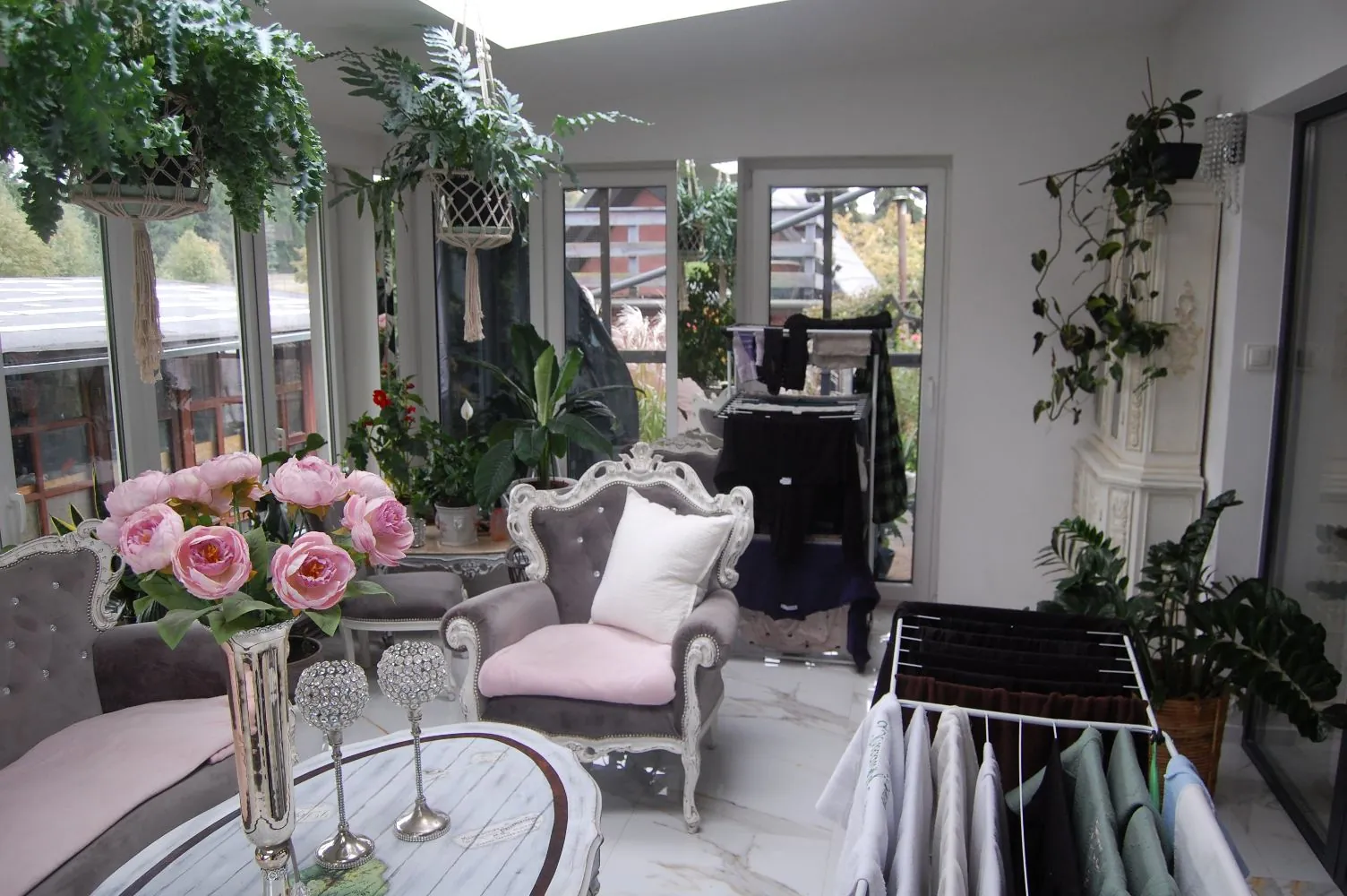 Wnętrze jasnego pomieszczenia przypominającego oranżerię lub butik. W centrum znajduje się szary, ozdobny fotel z różową poduszką i białką poduszką opartą o oparcie. Po lewej stronie widoczny jest fragment szarej kanapy z różowym kocem oraz stolik z marmurowym blatem, na którym stoi srebrny wazon z różowymi piwoniami i dwa ozdobne świeczniki. W tle widać wieszaki z ciemną odzieżą oraz duże okna z widokiem na zieleń. W pomieszczeniu znajduje się wiele roślin doniczkowych, w tym rośliny zwisające z sufitu oraz duże liściaste rośliny w donicach na podłodze i półkach.