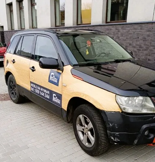 Czarny samochód typu SUV zaparkowany na kostce brukowej przed budynkiem. Na boku pojazdu znajduje się okleina imitująca jasne drewno z niebieskim logo 'MEBLE FERENS' oraz napisem 'MEBLE NA WYMIAR tel.: 509 144 244'.