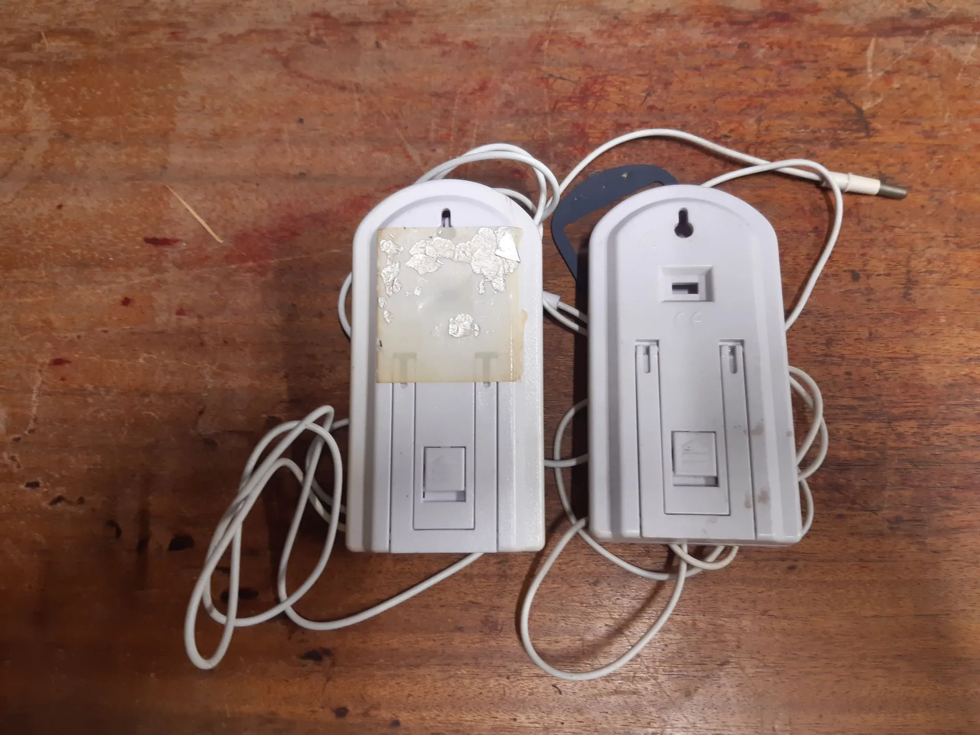 Dwa białe, plastikowe urządzenia elektroniczne o zaokrąglonej górnej krawędzi, leżące obok siebie na brązowej, drewnianej powierzchni. Oba urządzenia mają podłączone białe kable. Lewe urządzenie posiada na przodzie resztki odklejonej etykiety. Prawe urządzenie ma widoczne małe wgłębienie i oznaczenie CE. Jeden z kabli kończy się wtykiem typu jack.