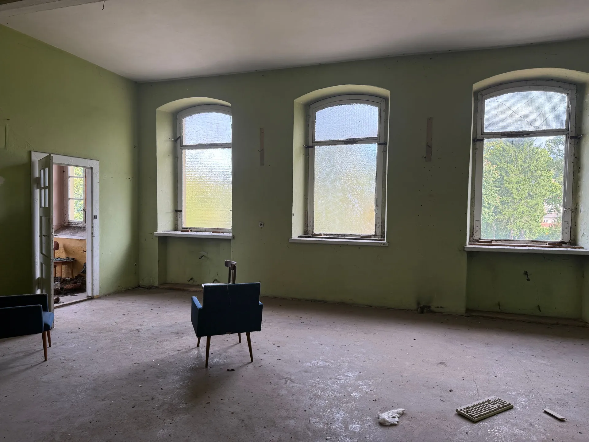 Wnętrze pustego, zaniedbanego pomieszczenia z jasnozielonymi ścianami i betonową podłogą. W ścianie znajdują się trzy duże, łukowate okna z białymi ramami, przez które wpada światło i widać zieleń drzew. Po lewej stronie widoczne są otwarte drzwi prowadzące do innego pomieszczenia. W centrum pokoju stoi pojedynczy, ciemnoniebieski fotel skierowany tyłem do widza, a w lewym dolnym rogu fragment drugiego podobnego fotela. Na podłodze leżą drobne odpady i kawałek białej kratki.