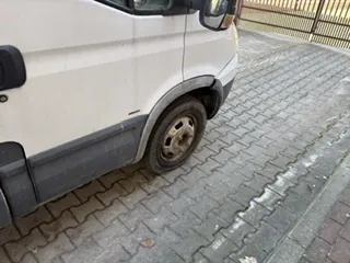 Biały furgon zaparkowany na brukowanej nawierzchni. Widoczna przednia część pojazdu z kołem, brudnymi plamami na boku i lusterkiem bocznym. W tle metalowe ogrodzenie. Nawierzchnia złożona z kamiennych płyt ułożonych w wzór herringbone, częściowo mokra. Brak czytelnego tekstu.