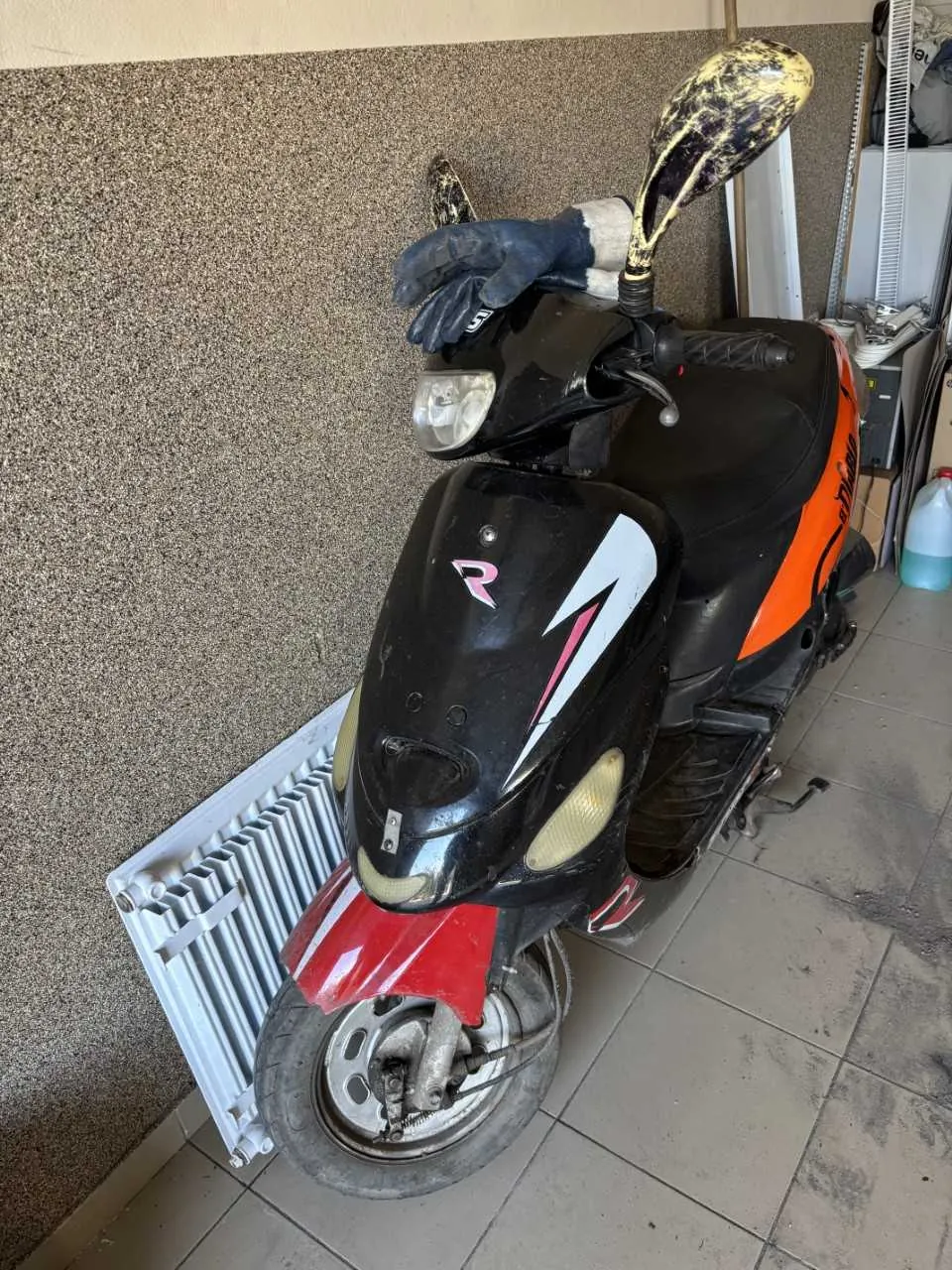 Czarny skuter z pomarańczowymi i czerwonymi akcentami, z różowym logo "R" i białą błyskawicą na przodzie. Na kierownicy leży czarna rękawica z białym mankietem. Skuter stoi w pomieszczeniu z teksturowaną ścianą, białym grzejnikiem i płytkami na podłodze. W tle widoczne są narzędzia oraz pojemnik z niebieską cieczą.