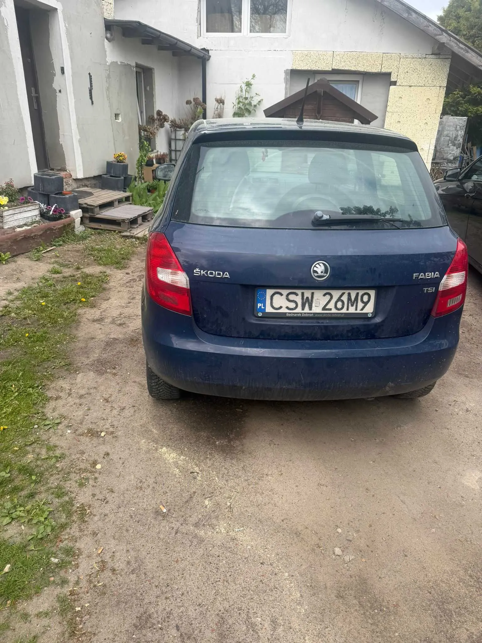 Z tyłu niebieski samochód Škoda Fabia TSI z tablicą rejestracyjną CSW 26M9 (PL). Samochód stoi na podwórku przed białym domem z żółtymi elementami elewacji. Po lewej stronie widoczne są donice z kwiatami, stosy opon i drewniane palety. Na ziemi brudna droga z plamami trawy. W tle dom z oknami i roślinami.
