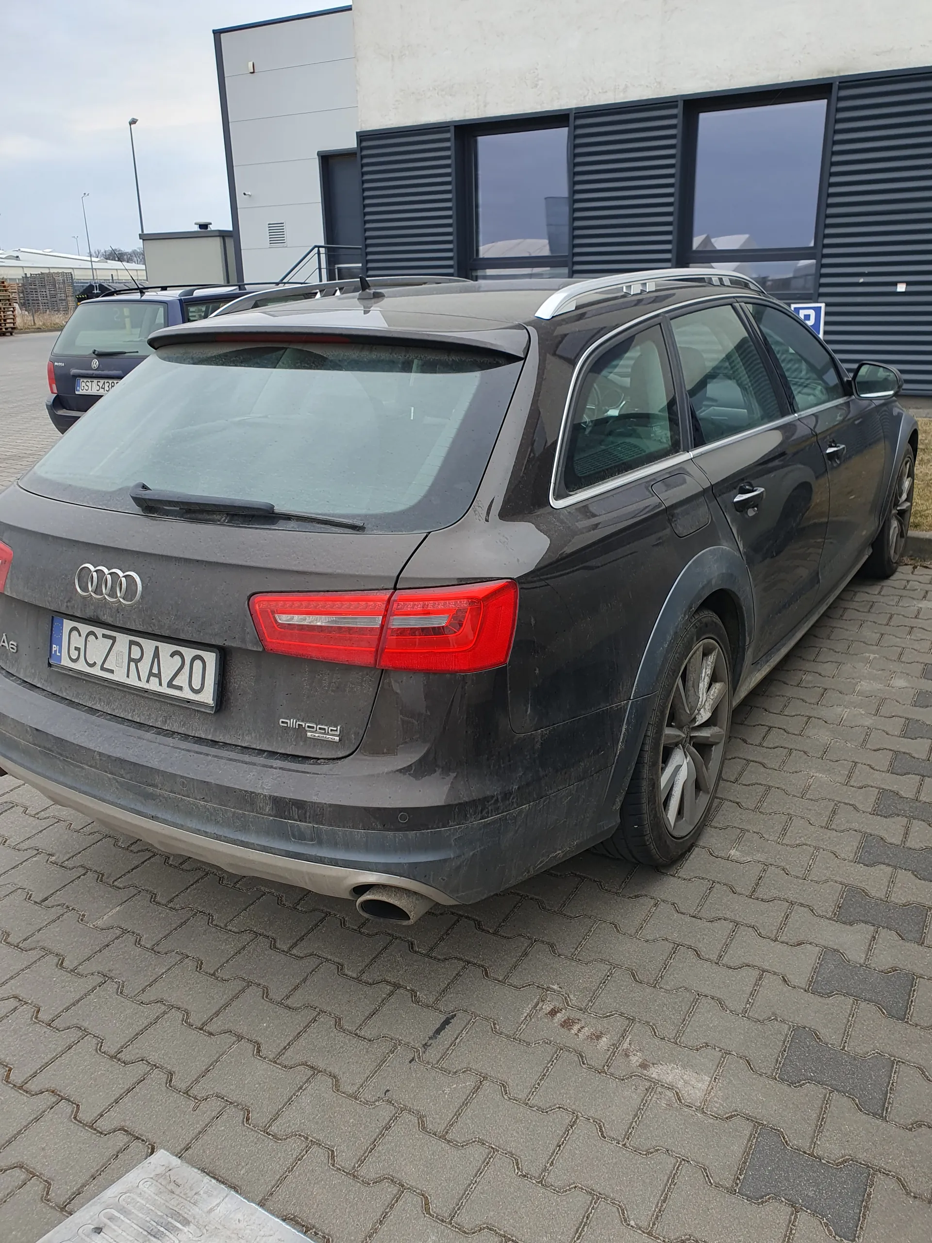 Ciemnoszary samochód Audi typu kombi zaparkowany na brukowanej powierzchni. Tablica rejestracyjna z napisem GCZ RA20. W tle budynki z dużymi oknami i nowoczesnym wykończeniem oraz drugi samochód.