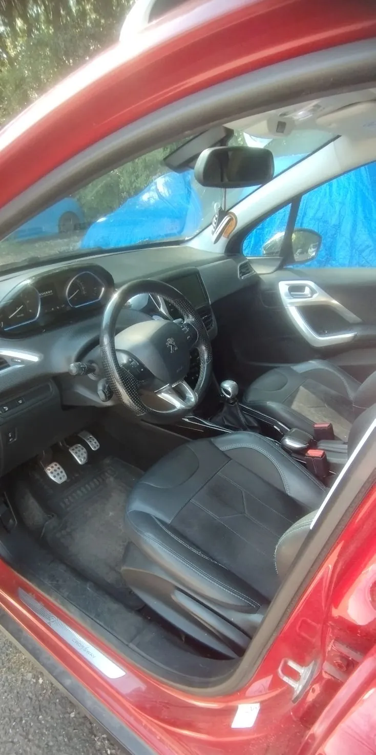 Wnętrze czerwonego samochodu widziane z perspektywy otwartych drzwi kierowcy. Widoczny jest czarny fotel z materiału, kierownica z logo marki Peugeot, deska rozdzielcza z zegarami, pedały oraz lewarek zmiany biegów. Przez przednią szybę i lusterko wsteczne widać niebieską plandekę w tle.