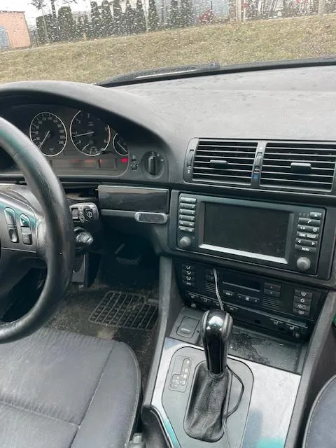 Wnętrze samochodu z widokiem na deskę rozdzielczą, kierownicę po lewej stronie, wskaźniki prędkości i obrotomierza, centralną konsolę z systemem audio, przyciskami i dźwignią zmiany biegów. Widoczne są również siedzenia oraz zewnętrzny krajobraz za przednią szybą z trawą i drzewami.